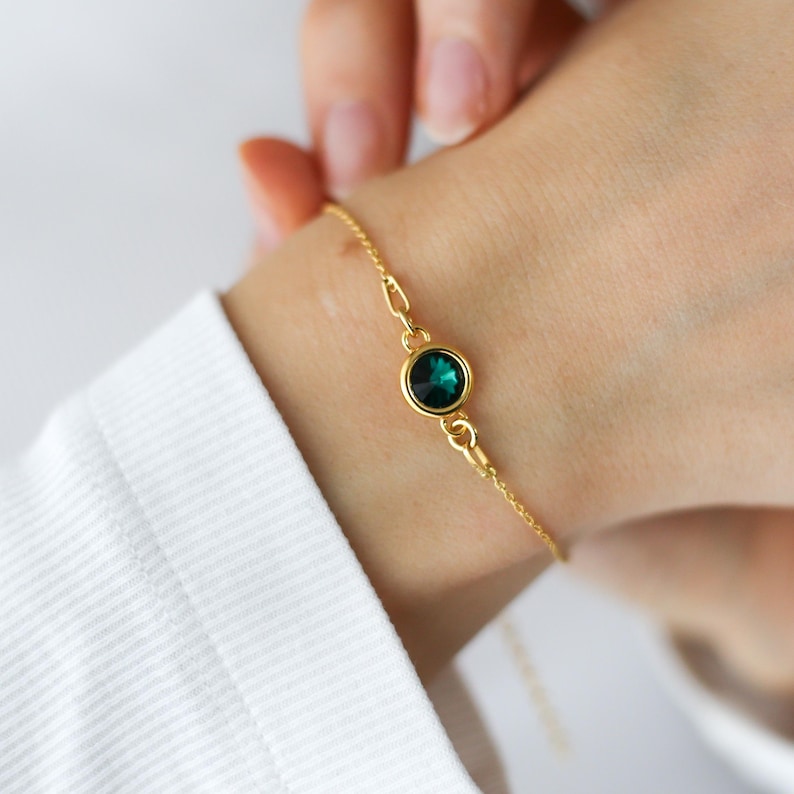 May include: Gold-toned bracelet featuring a round, emerald-green gemstone. The delicate chain is attached to a bezel setting that holds the gemstone. The bracelet is worn on a wrist, with a white shirt in the background.