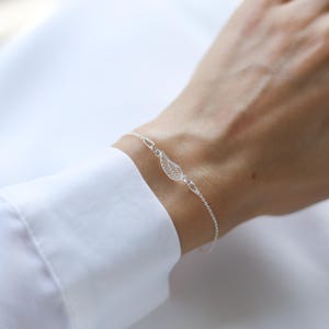 May include: A delicate silver bracelet featuring a detailed wing charm. The bracelet is worn on a wrist, with a white shirt in the background. The wing charm is the focal point, showcasing intricate feather detailing.