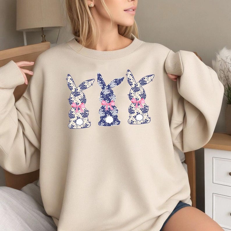 Puede incluir: Sudadera beige con tres dise&ntilde;os de conejos. Cada conejo tiene un estampado floral azul y un lazo rosa. La sudadera es de color neutro, adecuada para ropa informal.