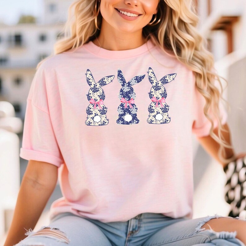 Puede incluir: Camiseta rosa claro con tres dise&ntilde;os de conejos. Los conejos tienen un estampado floral azul, lazos rosas y colas blancas. La camiseta tiene mangas cortas y un corte holgado.