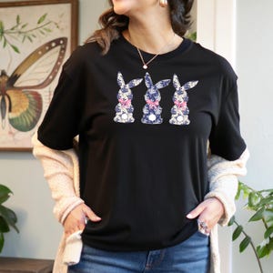 Puede incluir: Camiseta negra con tres dise&ntilde;os de conejos. Cada conejo est&aacute; estampado con estampados florales azules y blancos y un lazo rosa. Los conejos est&aacute;n dispuestos en una fila en la parte delantera de la camiseta, creando un dise&ntilde;o primaveral.