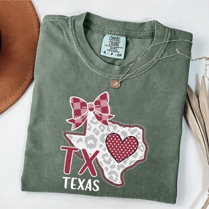 May include: An olive green t-shirt with a graphic of the state of Texas. The design includes a red and white plaid bow, a leopard print pattern, and a red polka dot heart. The text "TX Texas" is printed below.