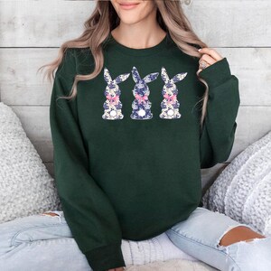Puede incluir: Una sudadera verde oscuro con tres dise&ntilde;os de conejos florales. Cada conejo tiene un estampado floral azul, blanco y rosa, y un lazo rosa. La sudadera es de manga larga y cuello redondo.