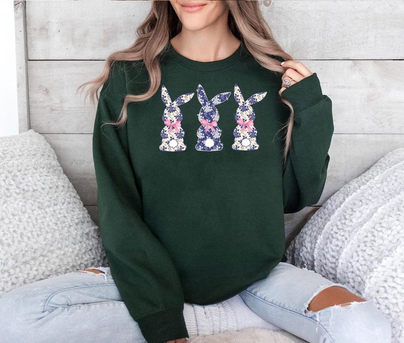 Puede incluir: Una sudadera verde oscuro con tres dise&ntilde;os de conejos florales. Cada conejo tiene un estampado floral azul, blanco y rosa, y un lazo rosa. La sudadera es de manga larga y cuello redondo.