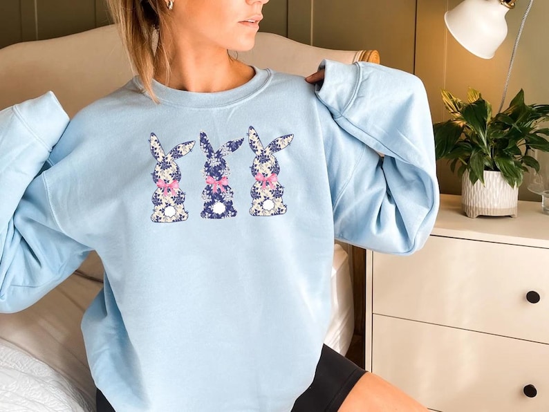Puede incluir: Sudadera azul claro con tres dise&ntilde;os de conejos. Cada conejo tiene un estampado floral en blanco, azul y rosa, con un lazo rosa. La sudadera es de cuello redondo.