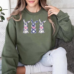 Puede incluir: Una sudadera verde oliva con tres dise&ntilde;os de conejos. Cada conejo tiene un estampado floral en azul, blanco y rosa, con un lazo rosa. La sudadera es una parte superior informal de manga larga.