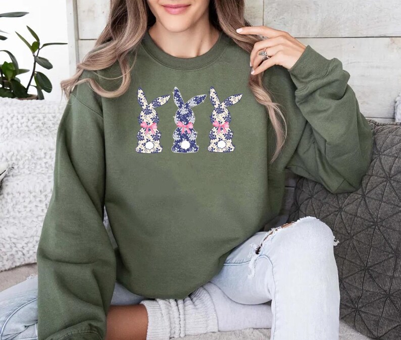 Puede incluir: Una sudadera verde oliva con tres dise&ntilde;os de conejos. Cada conejo tiene un estampado floral en azul, blanco y rosa, con un lazo rosa. La sudadera es una parte superior informal de manga larga.