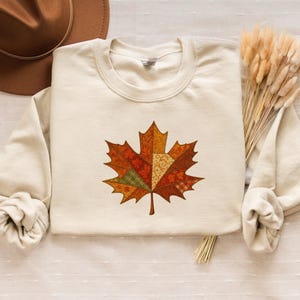 Peut inclure: Sweat-shirt crème avec un motif de feuille d'érable colorée. La feuille est composée de couleurs automnales, orange, rouge et vert, avec un motif patchwork. Un chapeau marron et des épis de blé séchés sont également visibles.