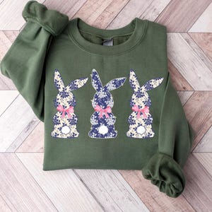 Puede incluir: Una sudadera verde oliva con tres dise&ntilde;os de conejos. Cada conejo tiene un estampado floral en azul y blanco, con un lazo rosa. La sudadera tiene mangas largas y cuello redondo.