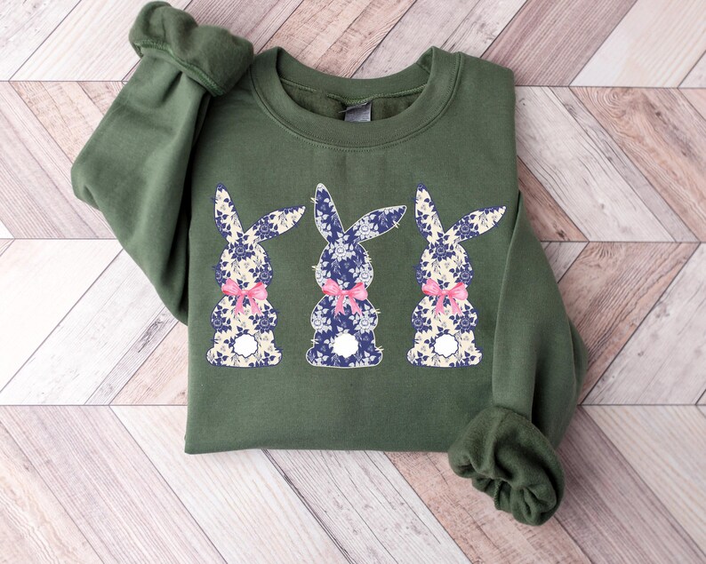 Puede incluir: Una sudadera verde oliva con tres dise&ntilde;os de conejos. Cada conejo tiene un estampado floral en azul y blanco, con un lazo rosa. La sudadera tiene mangas largas y cuello redondo.