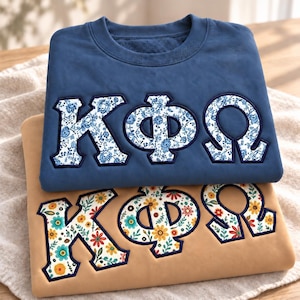 May include: A blue crewneck sweatshirt featuring Greek letters in a blue and white floral pattern, layered over a tan sweatshirt with the same Greek letters in a colorful floral print with red, yellow, and teal flowers.