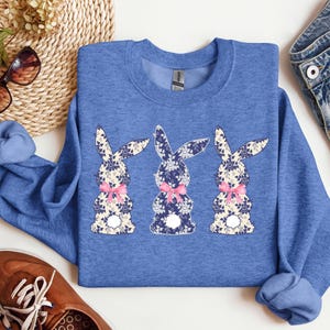 Puede incluir: Una sudadera azul con cuello redondo con tres dise&ntilde;os de conejos florales. Cada conejo tiene un estampado floral azul, blanco y beige, con un lazo rosa. La sudadera es de color azul liso.