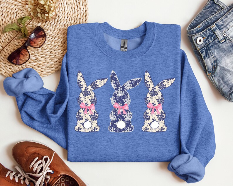 Puede incluir: Una sudadera azul con cuello redondo con tres dise&ntilde;os de conejos florales. Cada conejo tiene un estampado floral azul, blanco y beige, con un lazo rosa. La sudadera es de color azul liso.