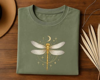 Puede incluir: Camiseta verde oliva con un diseño de libélula dorada, una luna creciente y estrellas. La libélula tiene alas y cuerpo detallados. La camiseta está doblada sobre una superficie de madera, con un sombrero y un abanico de hojas decorativas en el fondo.