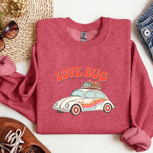 May include: A red sweatshirt with the words "LOVE BUG" in orange lettering above a white car with a rainbow stripe and flowers on top. The sweatshirt has a crew neck and long sleeves.