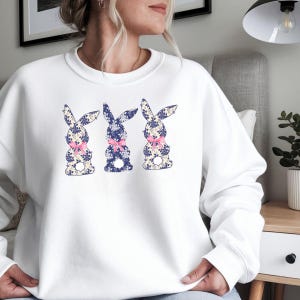 Puede incluir: Sudadera blanca con tres dise&ntilde;os de conejos. Cada conejo tiene un estampado floral en azul y crema, con un lazo rosa. La sudadera es de cuello redondo.