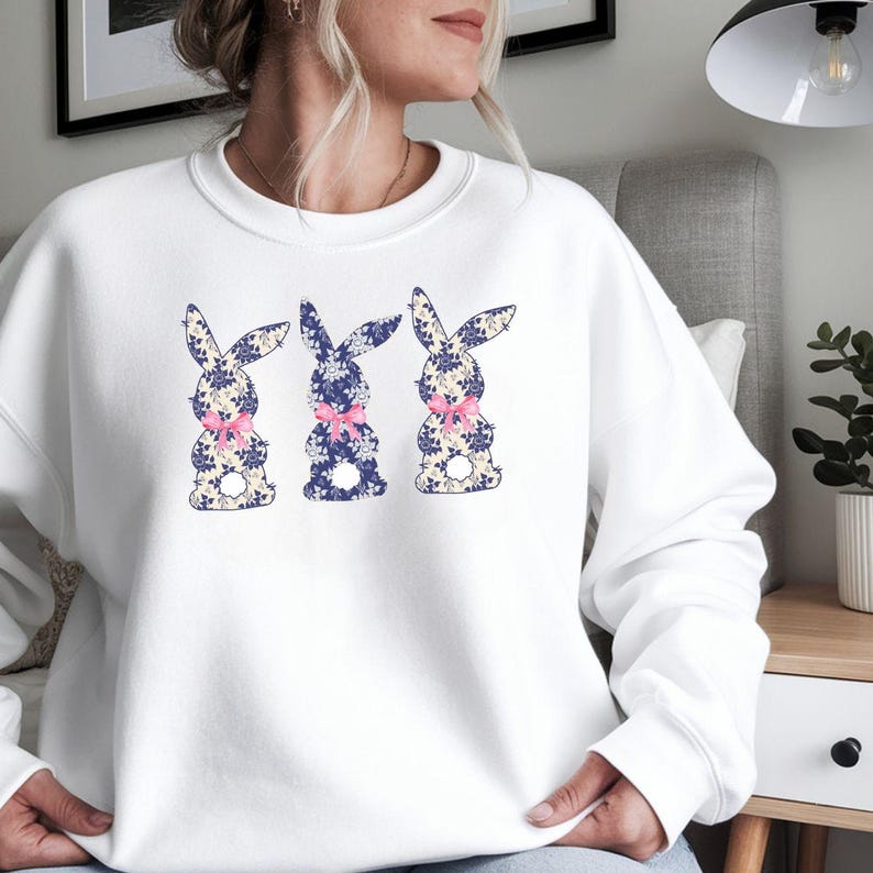 Puede incluir: Sudadera blanca con tres dise&ntilde;os de conejos. Cada conejo tiene un estampado floral en azul y crema, con un lazo rosa. La sudadera es de cuello redondo.