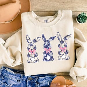 Puede incluir: Sudadera color crema con tres siluetas de conejos con estampados florales azules y lazos rosas. Los conejos est&aacute;n centrados en la parte delantera de la parte superior de manga larga. La sudadera se combina con pantalones cortos de mezclilla y un sombrero marr&oacute;n.