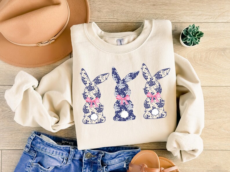 Puede incluir: Sudadera color crema con tres siluetas de conejos con estampados florales azules y lazos rosas. Los conejos est&aacute;n centrados en la parte delantera de la parte superior de manga larga. La sudadera se combina con pantalones cortos de mezclilla y un sombrero marr&oacute;n.