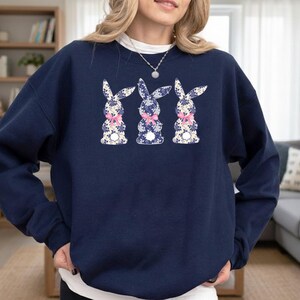Puede incluir: Sudadera azul marino con tres conejos con estampados florales y lazos rosas. Los conejos est&aacute;n centrados en la parte delantera de la sudadera. Cuello redondo y mangas largas.