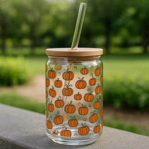 May include: A clear glass tumbler with a bamboo lid and a glass straw. The tumbler is decorated with orange pumpkin illustrations. The background is a blurred green outdoor scene.