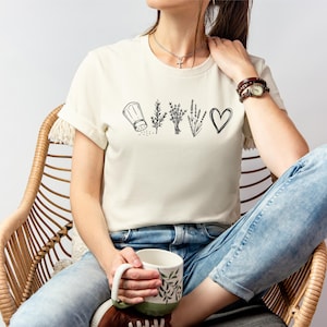 May include: A cream-coloured short-sleeve t-shirt featuring a black line art graphic across the chest. The design displays a salt shaker, a sprig of rosemary, a bundle of herbs, a sprig of lavender, and a heart symbol arranged in a horizontal row.