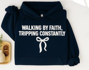 May include: Navy blue hoodie with the text "WALKING BY FAITH, TRIPPING CONSTANTLY" in white block letters. A white bow graphic is below the text. The hoodie has a front pocket and a hood.