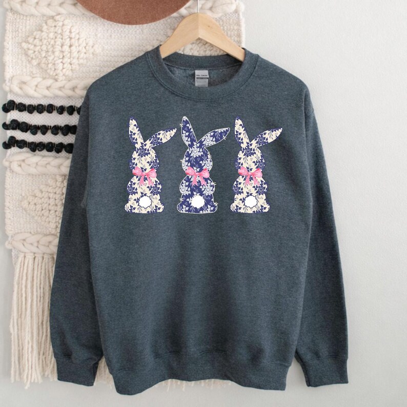 Puede incluir: Una sudadera gris oscuro con tres dise&ntilde;os de conejos. Cada conejo tiene un estampado floral en azul y blanco, con un lazo rosa. La sudadera est&aacute; colgada en una percha de madera.