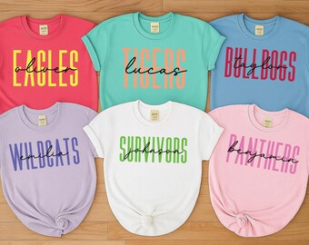 May include: Six colorful t-shirts with team names and individual names printed on them. The shirts are in various colors, including red, teal, blue, purple, white, and pink. The text is in bold, block letters with a cursive name underneath.