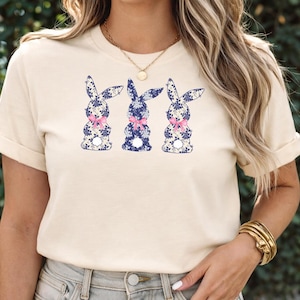 Puede incluir: Camiseta color crema con tres siluetas de conejos. Cada conejo est&aacute; decorado con un estampado floral azul y un lazo rosa. Los conejos est&aacute;n dispuestos en fila en la parte delantera de la camiseta.