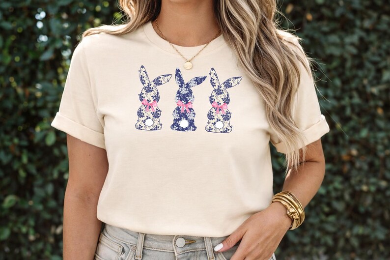 Puede incluir: Camiseta color crema con tres siluetas de conejos. Cada conejo est&aacute; decorado con un estampado floral azul y un lazo rosa. Los conejos est&aacute;n dispuestos en fila en la parte delantera de la camiseta.