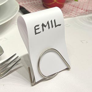 Könnte beinhalten: Eine weiße Tischkarte mit dem Namen "EMIL" in schwarzer Schrift, gehalten von einem silbernen Metallhalter. Die Karte liegt auf einer weißen Tischdecke, mit Besteck und einer dekorativen Pflanze im Hintergrund.