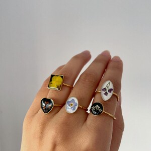 Puede incluir: Una mano muestra seis anillos dorados, cada uno con una flor seca diferente encapsulada en resina. Los anillos tienen varias formas, incluyendo cuadrada, ovalada y de corazón, con flores en amarillo, azul y morado.