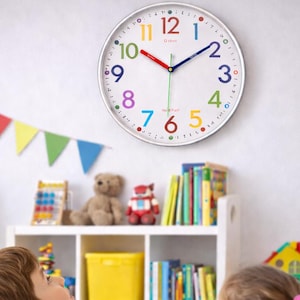 Könnte beinhalten: Eine weiße Wanduhr mit bunten Zahlen und Zeigern hängt über einem Tisch, an dem zwei Kinder mit Bausteinen spielen. Ein farbenfrohes Banner und ein Regal mit Spielzeug befinden sich im Hintergrund.
