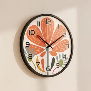 Puede incluir: Un reloj de pared redondo con marco negro. La esfera presenta un gran diseño de flor naranja, manecillas negras y números negros. También hay elementos florales adicionales en azul, verde y amarillo.