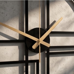 May include: A large circular wall clock featuring a black metal frame with a geometric grid pattern. The center consists of a solid black circular hub with three slender gold-colored hands indicating the time against a textured light gray background.