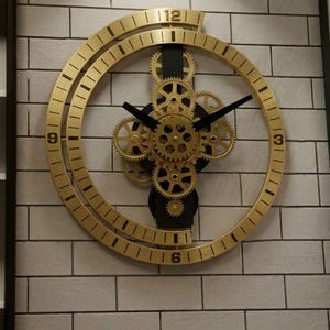Op de afbeelding: Een goudkleurige, ronde wandklok met zichtbare tandwielen en zwarte wijzers. De wijzerplaat heeft Romeinse cijfers en is gemonteerd op een witte bakstenen muur. Het ontwerp van de klok bevat een steampunk-esthetiek.