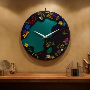 Puede incluir: Un reloj de pared redondo con un fondo oscuro y una forma verde azulado en el centro. La esfera del reloj est&aacute; decorada con flores secas de colores. Las manecillas blancas indican la hora. El reloj est&aacute; colgado en una pared beige.
