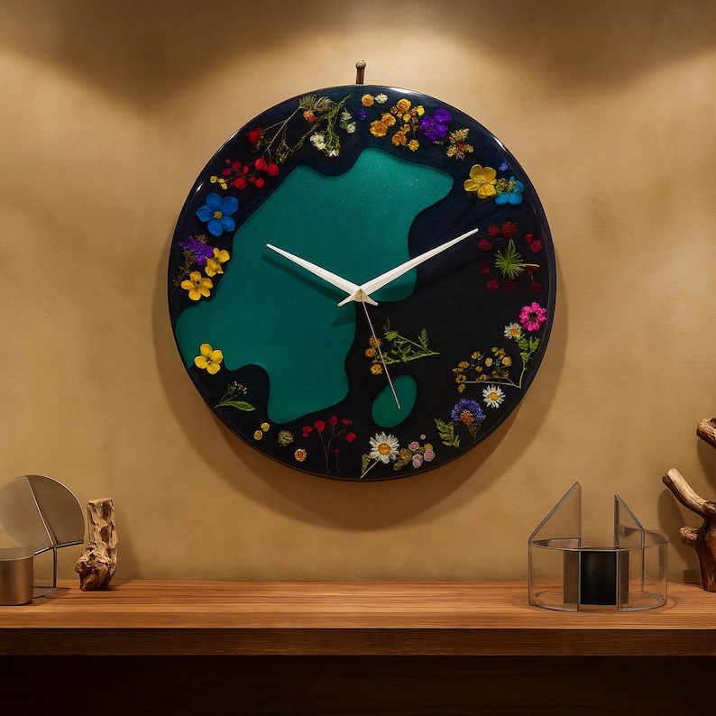 Puede incluir: Un reloj de pared redondo con un fondo oscuro y una forma verde azulado en el centro. La esfera del reloj est&aacute; decorada con flores secas de colores. Las manecillas blancas indican la hora. El reloj est&aacute; colgado en una pared beige.