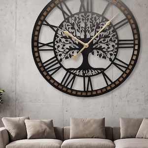 Può includere: Un grande orologio da parete circolare con una cornice in metallo nero e una silhouette centrale dell'albero della vita. Il quadrante mostra numeri romani e due sottili lancette color oro. Un bordo marrone testurizzato si trova dietro i numeri.