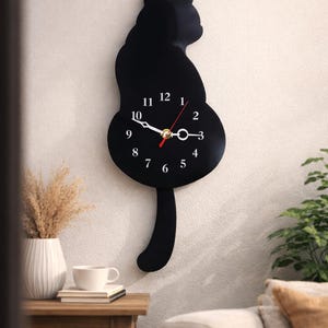 Pode incluir: Um relógio de parede preto em forma de gato com números e ponteiros brancos. O design do relógio inclui uma cauda e um corpo circular, com o mostrador integrado no corpo. O relógio está montado numa parede branca.
