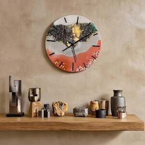 Puede incluir: Un reloj de pared redondo con un dise&ntilde;o &uacute;nico. La esfera del reloj presenta una mezcla de colores que incluyen negro, dorado, blanco, gris y coral. Las manecillas y los marcadores negros indican la hora. El reloj est&aacute; montado en una pared beige.