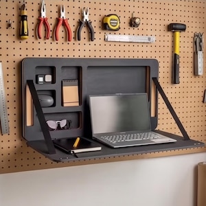 May include: A dark gray, wall-mounted desk with a laptop, notebook, and accessories. Tools like pliers, a hammer, and a tape measure are mounted on the pegboard above. The desk has a fold-down design for space-saving functionality.