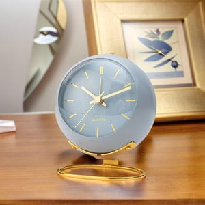 Può includere: Un orologio da tavolo al quarzo rotondo, di colore azzurro con una finitura opaca liscia. Il quadrante presenta sottili indicatori dorati e lancette abbinate. È sostenuto da un supporto in filo metallico color oro lucido con base ovale.