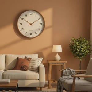 May include: A round wall clock with a light-coloured face, dark numerals, and thin hands. The clock features a wooden frame and is mounted on a solid-coloured wall above a sofa, a side table with a lamp, and a potted plant.