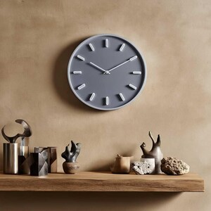 Puede incluir: Un reloj de pared redondo gris con manecillas y marcadores de hora blancos. El reloj est&aacute; montado en una pared beige sobre una estanter&iacute;a de madera con objetos decorativos. El di&aacute;metro del reloj es de aproximadamente 30 cm.