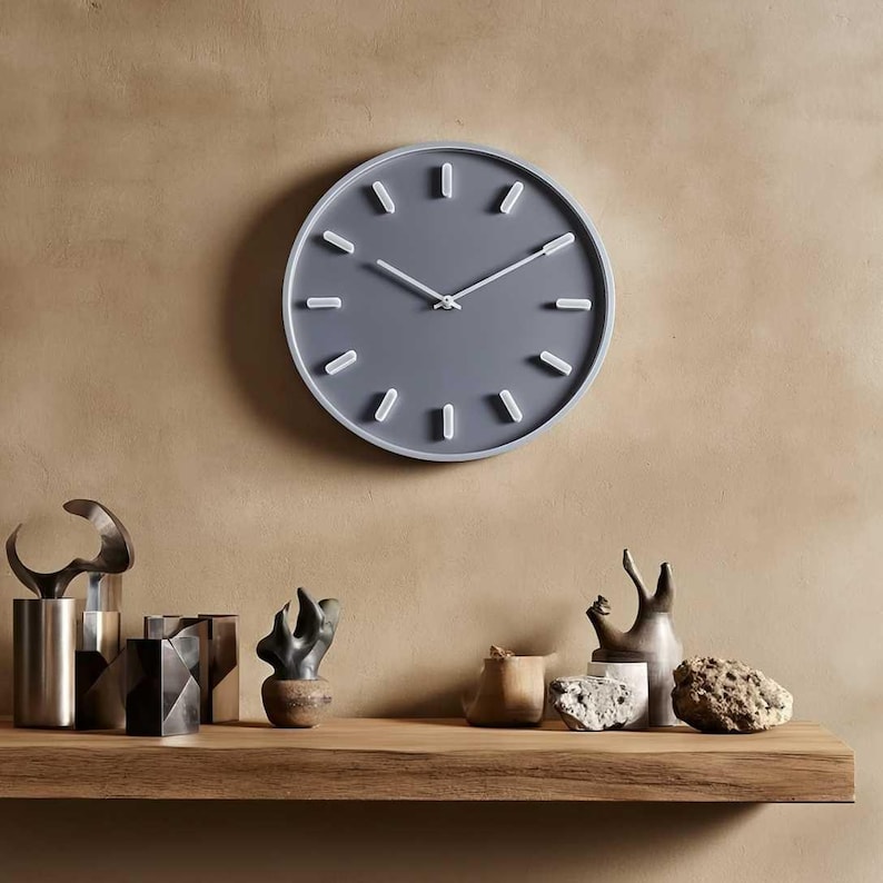 Puede incluir: Un reloj de pared redondo gris con manecillas y marcadores de hora blancos. El reloj est&aacute; montado en una pared beige sobre una estanter&iacute;a de madera con objetos decorativos. El di&aacute;metro del reloj es de aproximadamente 30 cm.