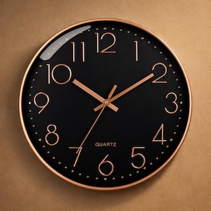 Op de afbeelding: Een ronde zwarte wandklok met een roségouden metalen frame. De wijzerplaat heeft roségouden cijfers en wijzers, met het woord "QUARTZ" onderaan. De klok heeft een modern design.