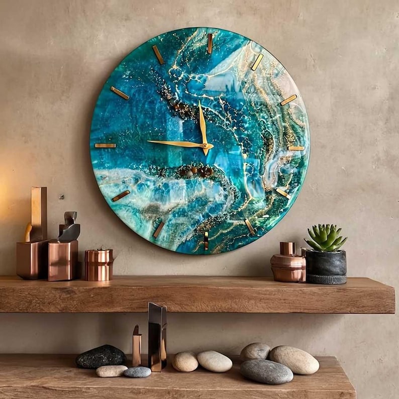 Puede incluir: Un reloj de pared redondo con un dise&ntilde;o inspirado en el oc&eacute;ano azul y verde. La esfera del reloj presenta manecillas e indicadores de hora dorados. El reloj est&aacute; montado en una pared de color neutro, con objetos decorativos en una estanter&iacute;a de madera.