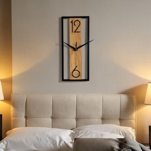 Pode incluir: Um relógio de parede retangular com moldura preta e centro de madeira clara. O mostrador do relógio apresenta os números 12 e 6 em preto, com ponteiros pretos. O relógio está montado numa parede de cor neutra acima de uma cama.